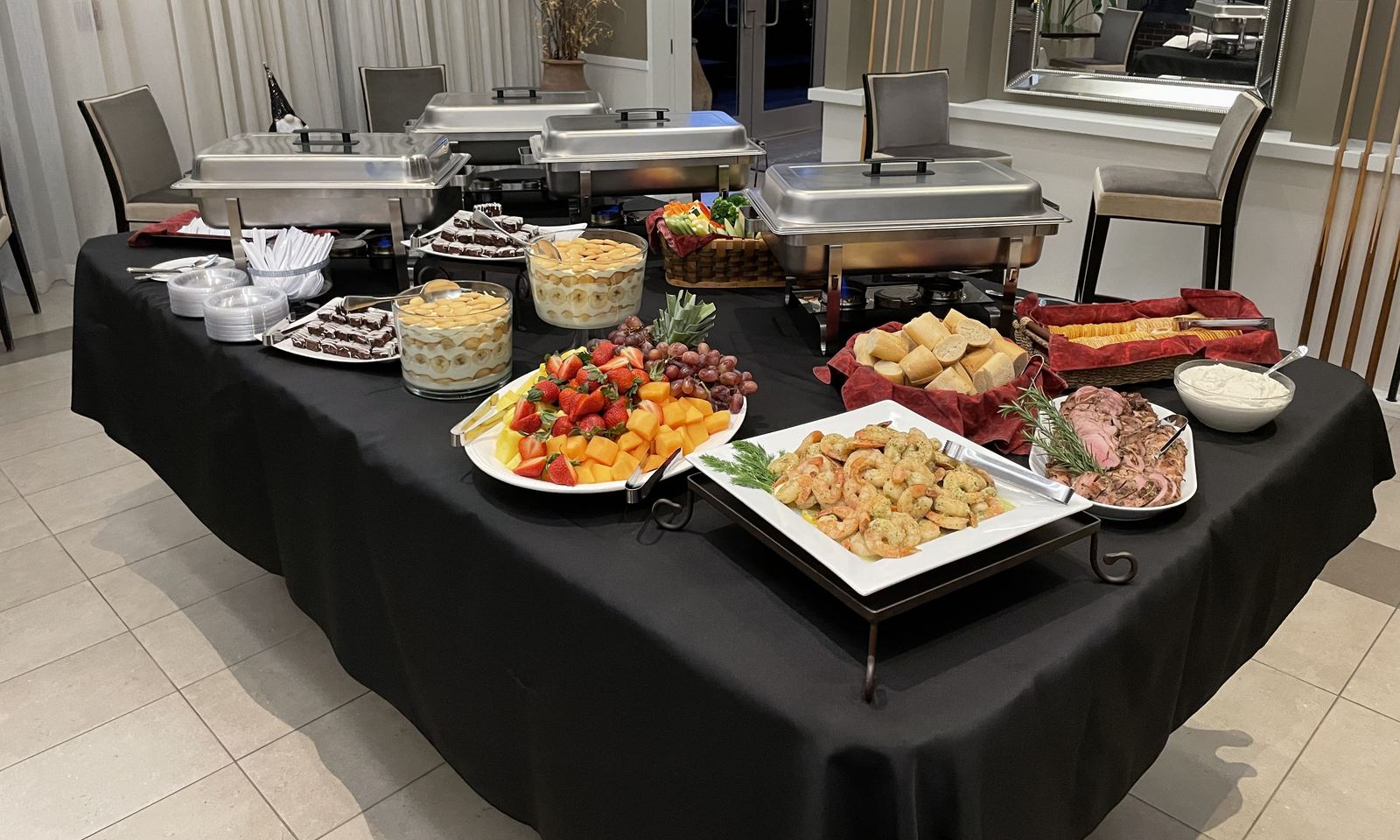 Catered event buffet with chafing dishes, a fresh fruit platter, shrimp and a basket of rolls on black linens