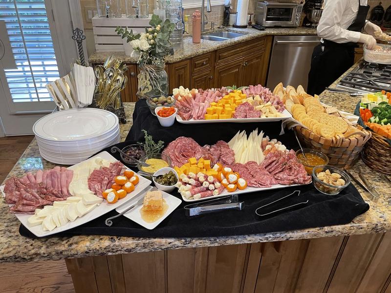 Large charcuterie spread of cured meats, cheeses, crackers and dips on a kitchen island with stacked plates and gold flatware