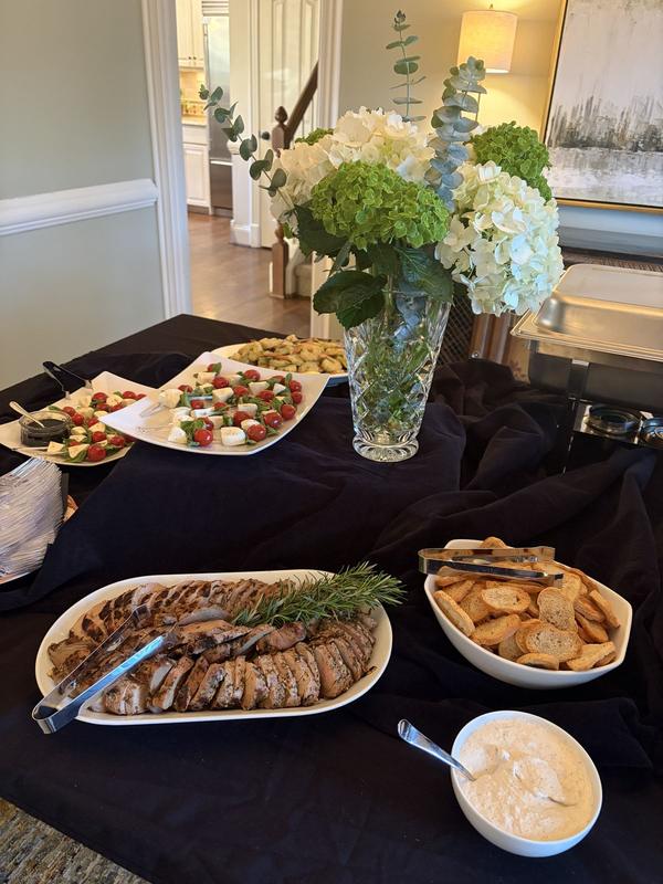 Party buffet with sliced roast garnished with rosemary, caprese skewers, crostini and a creamy dip beside a vase of hydrangeas