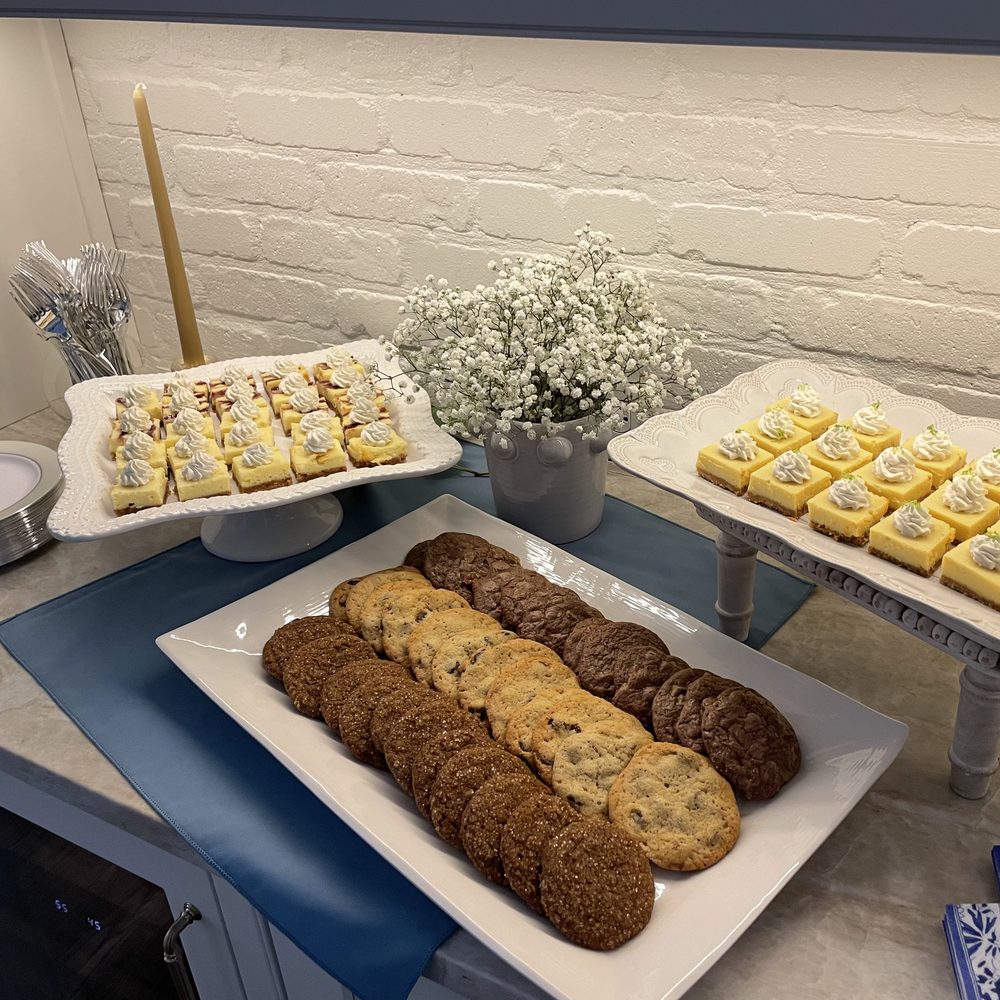 Dessert table with chocolate chip cookies and two raised trays of lemon and cheesecake-style bars beside baby’s breath